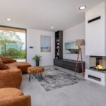 Wohnzimmer mit braunem Sofa, Kamin und Blick ins Grüne, geeignet für die Vermietung als Ferienobjekt durch Favorent.