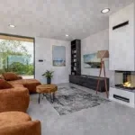 Wohnzimmer mit braunem Sofa, Kamin und Blick ins Grüne, geeignet für die Vermietung als Ferienobjekt durch Favorent.