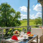 Gedeckter Tisch mit Blumenschale auf dem Balkon eines Ferienhauses mit Blick in die grüne Landschaft.