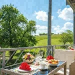 Gedeckter Tisch mit Blumenschale auf dem Balkon eines Ferienhauses mit Blick in die grüne Landschaft.