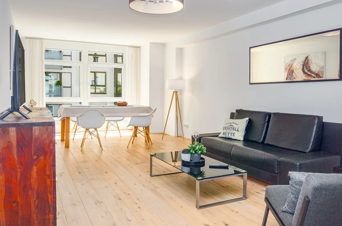 Wohnraum mit schwarzem Ledersofa, Glastisch und Essplatz vor großen Fenstern