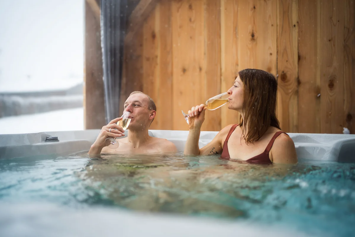 Paar mit Sektgläsern im Whirlpool vor Holzwand im Chalet Auszeit